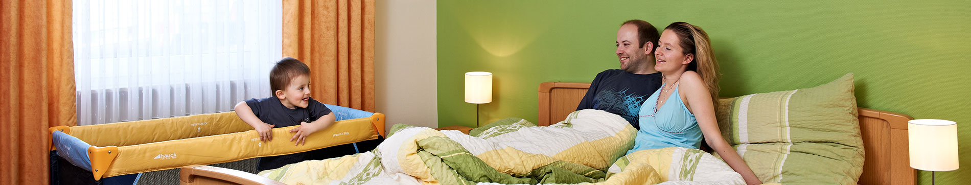Mutter und Vater die in einem Bett der Ferienwohnung sitzen und kleinem Sohn beim Spielen im Beistellbett zuschauen.
